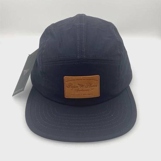 Spinning nylon navy Paper Planes 5 Panel Hat, leather logo on the front, with a white background.