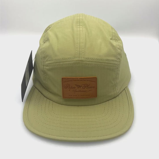 Spinning nylon olive green Paper Planes 5 Panel Hat, leather logo on the front,with a white background.