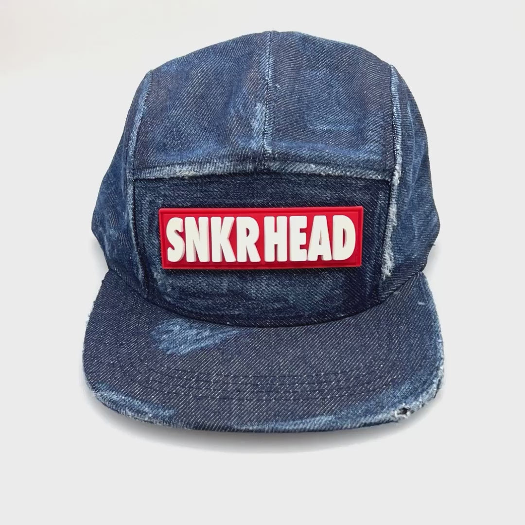 Spinning distressed blue denim SNKRHEAD 5 Panel Hat, rubber logo on the front, with a white background.