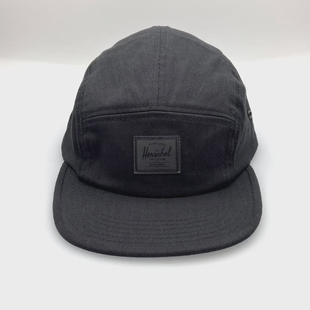 Spinning Black Herschel 5 Panel Hat with a black logo on the front panel, white background.