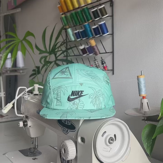 The making of a green NIKE Swim Shorts upcycled into a 5 Panel Hat, black iconic NIKE logo in front, with a white background.