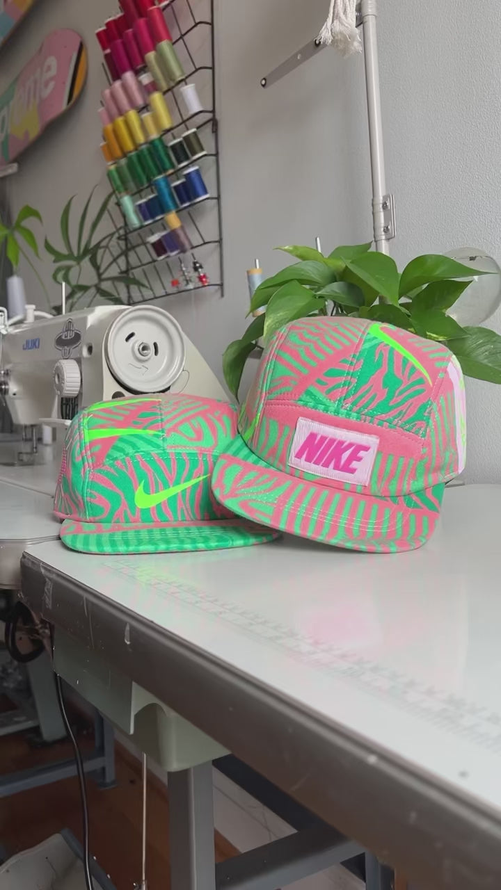 The making of a fluorescent green and pink Nike sweatshirt fleece upcycled into a 5 Panel Hat, Nike logos throughout, with a white background.