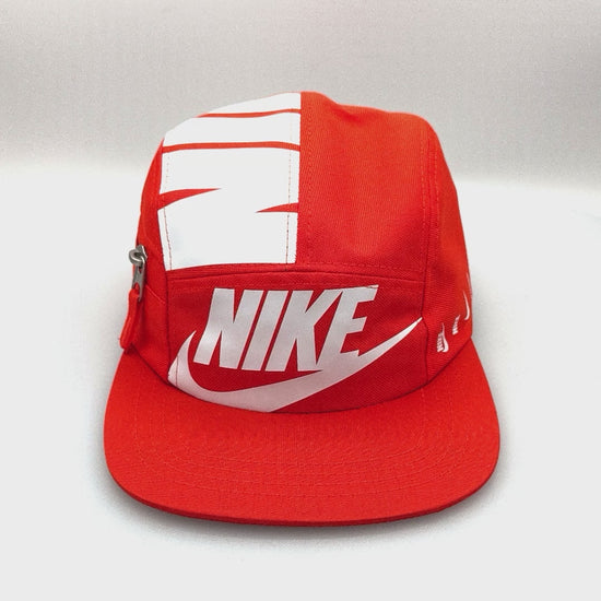 Spinning orange and white NIKE shoe box bag upcycled to a 5 Panel Hat, zipper on right side panel, with white background.