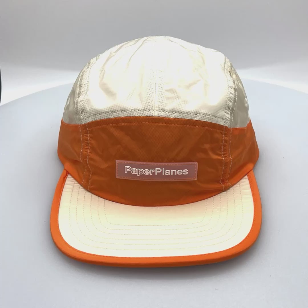 Spinning nylon orange and off white Paper Planes 5 Panel Hat, clear logo on the front, with a white background.