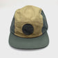 Spinning dark green and tan Mons Royale Alpine Adventure 5 panel hat, with white background.