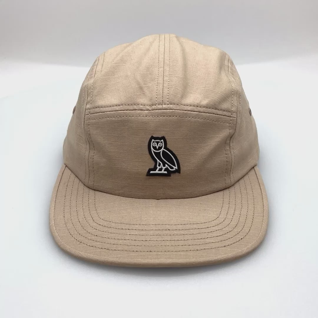 Spinning khaki OVO 5 Panel Hat, rubber OWL logo on front, with a white background.